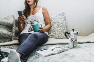 Woman relaxing on her bed holding a mug while looking at her phone.
