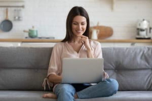 Smiling young woman sitting on her couch at home using laptop to research how to fix sagging breasts.