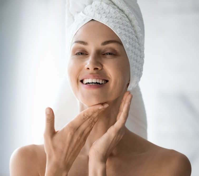 Happy woman with her hair wrapped in a towel looking at her in the bathroom mirror.