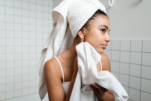 woman with hair wrapped in towel holding another towel to her face while looking in a mirror out of frame