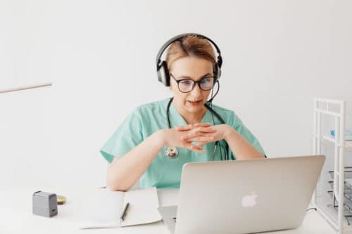 woman in blue scrubs wearing glasses and headset sitting in front of open laptop