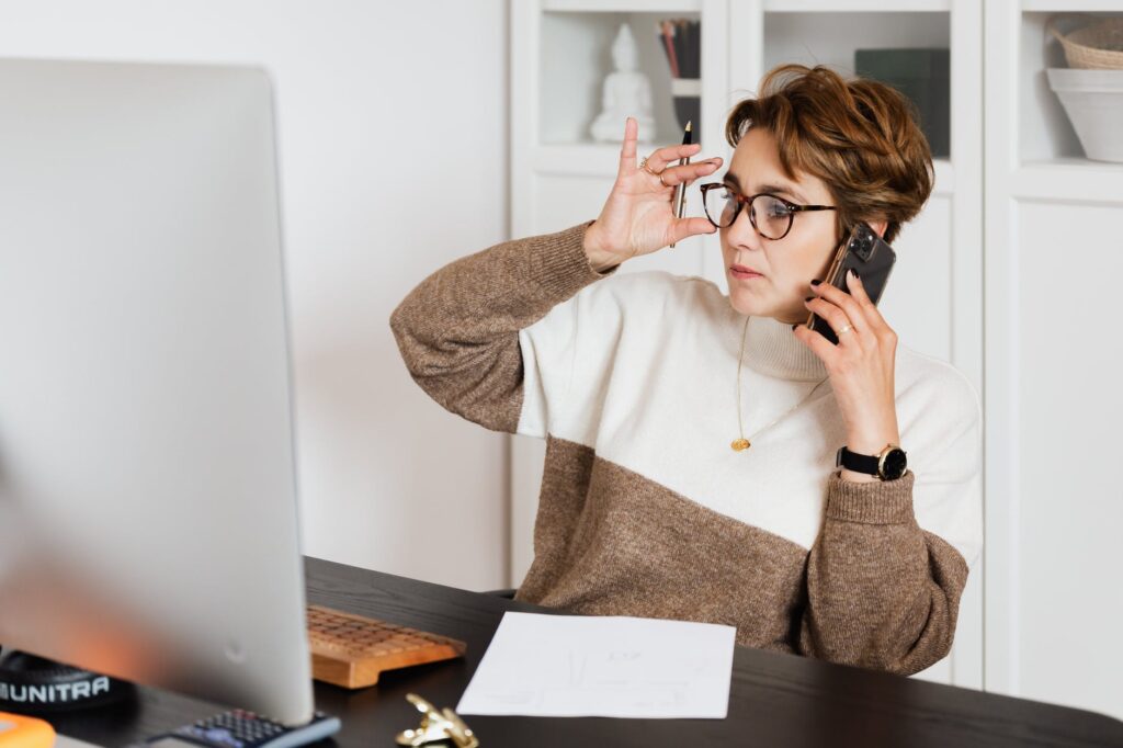 woman sitting at computer desk while holding phone up to her ear with other hand adjusting glasses